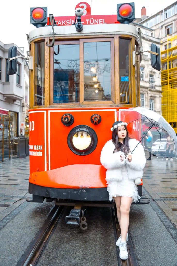 As the historic tram glides through the bustling pedestrian street, the presence of the Singaporean girl adds a touch of exotic elegance, creating a striking juxtaposition against the backdrop of Istanbul's historic charm.